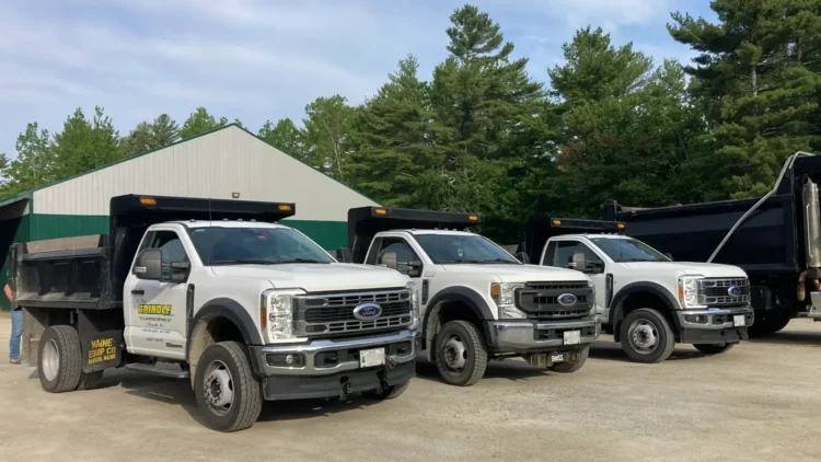 A fleet of white Ford Super Duty commercial dump trucks parked at a contractor's yard in Downeast Maine.