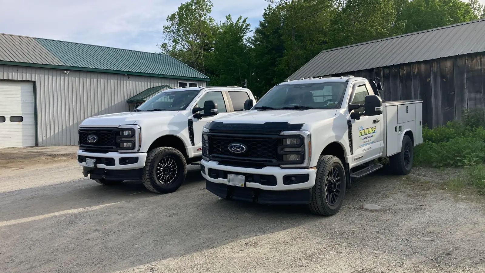 A detailed side-by-side view of two modern white Ford Super Duty Powerstroke diesel trucks from Grindle Landscaping of Ellsworth. On the right, an F-350 service truck with a specific utility body and the 'GRINDLE LANDSCAPING' logo clearly visible on the door. On the left, an F-350 crew cab pickup customized with intricate black multi-spoke wheels and aggressive oversized tires. They are parked on gravel in front of industrial buildings.