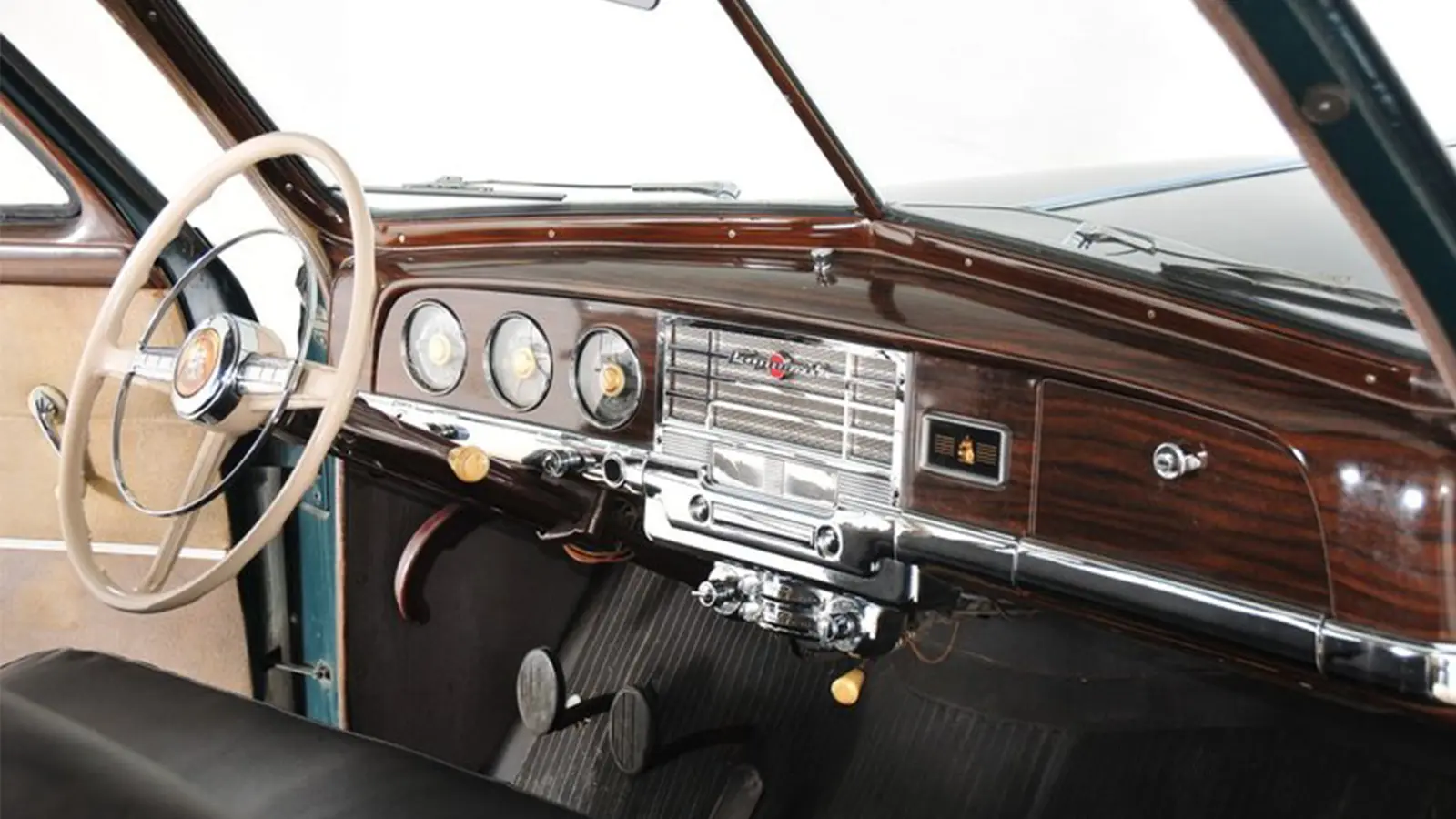 Interior view of a 1949 Plymouth showing the classic white steering wheel, chrome instrument panel, circular gauges, and signature faux woodgrain dashboard.