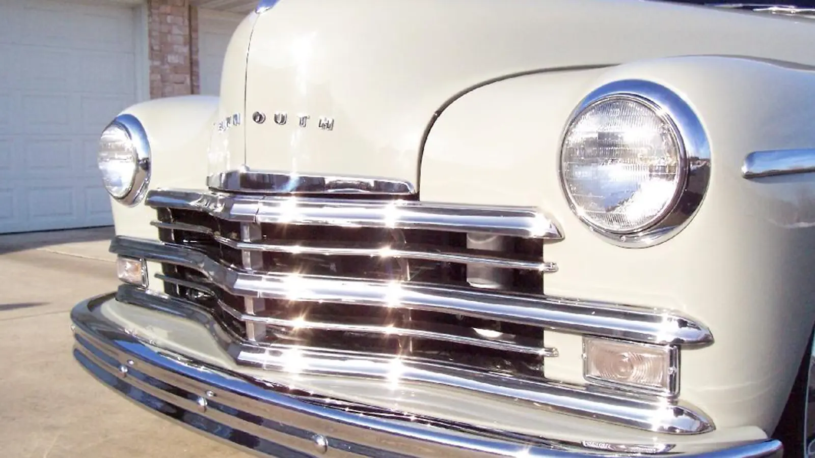 A close-up front angle of a light gray 1949 Plymouth Deluxe, highlighting the heavy chrome grille, passenger-side round headlight, and the distinct ribbed front bumper.