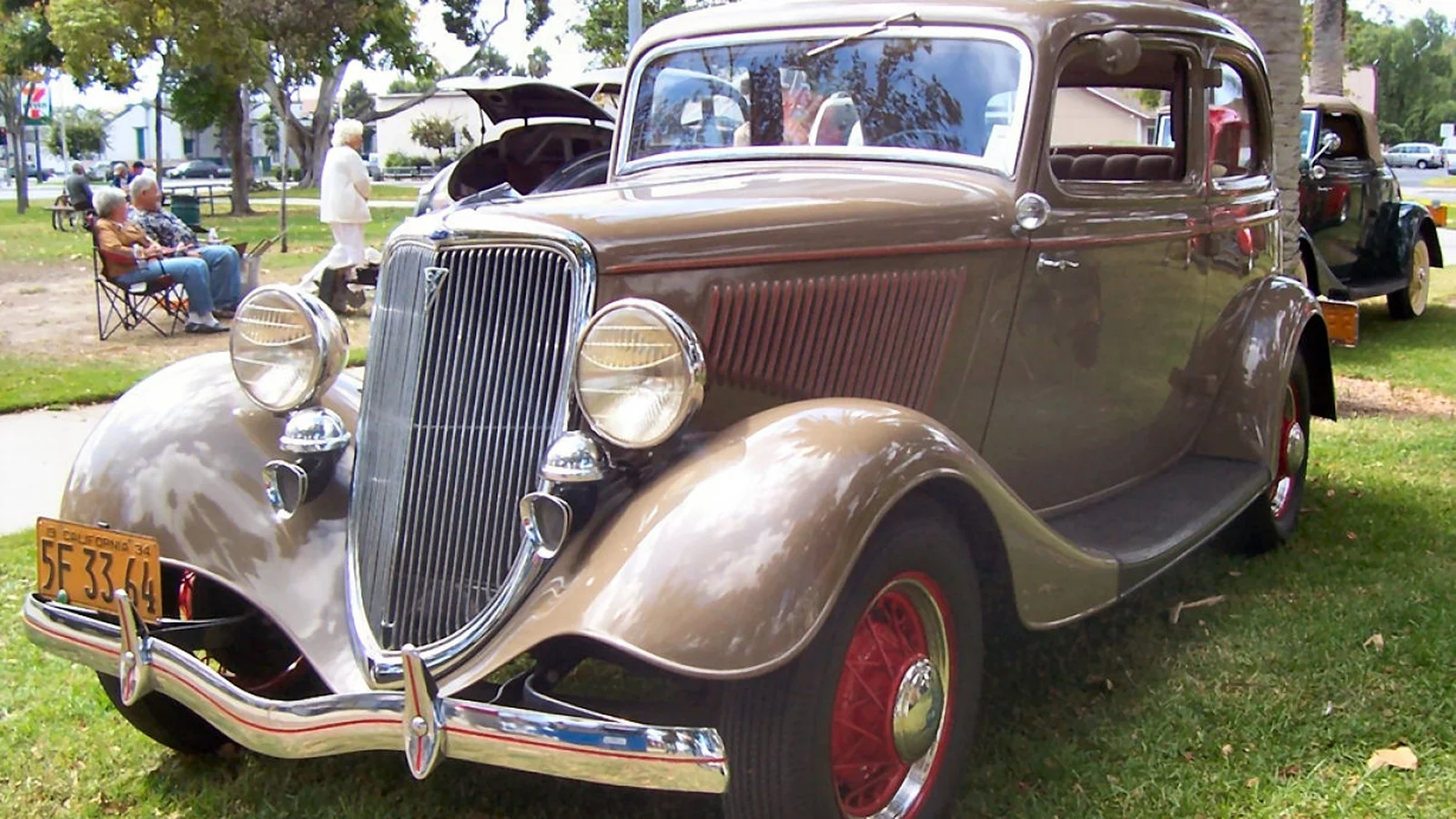 Restored tan 1934 Ford V8 with red wire wheels parked on grass at a classic car show