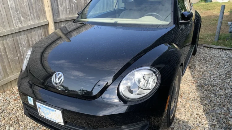 Black 2015 Volkswagen Beetle convertible parked on gravel by a wood fence, showing a Massachusetts Cape Cod and Islands license plate.