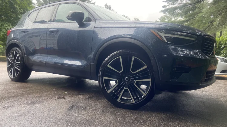 Dark blue Volvo XC40 compact SUV parked on a wet driveway lined with trees, front three-quarter view.