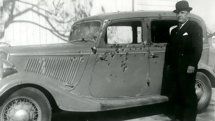 Black and white photo of Bonnie and Clyde's bullet-riddled 1934 Ford V8 sedan with a lawman in a suit and bowler hat standing beside it
