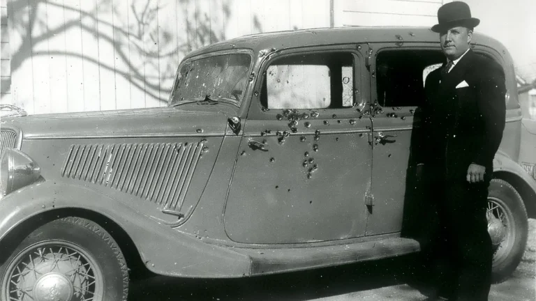 Black and white photo of Bonnie and Clyde's bullet-riddled 1934 Ford V8 sedan with a lawman in a suit and bowler hat standing beside it