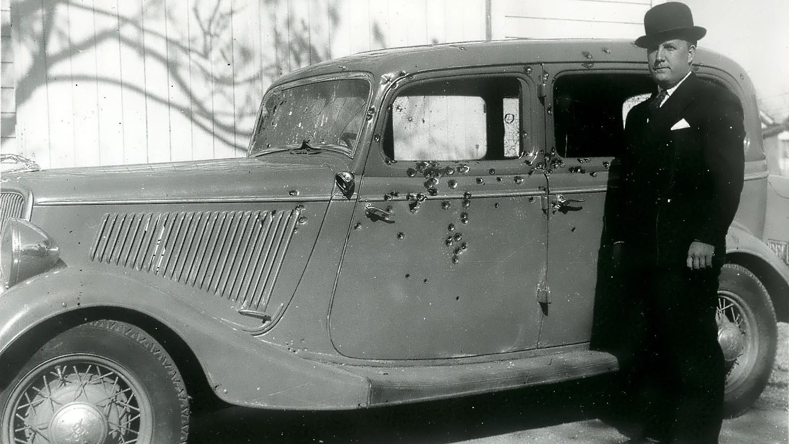 Black and white photo of Bonnie and Clyde's bullet-riddled 1934 Ford V8 sedan with a lawman in a suit and bowler hat standing beside it