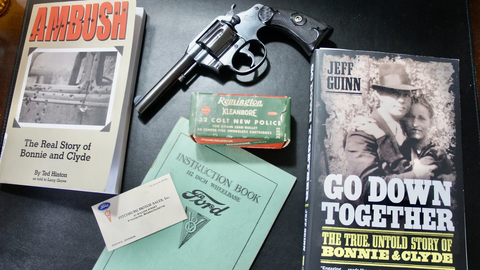 Bonnie and Clyde books, a Colt revolver, a Remington ammunition box, and a Ford V8 instruction book with a Fitchburg Motor Sales business card from Fitchburg, Massachusetts