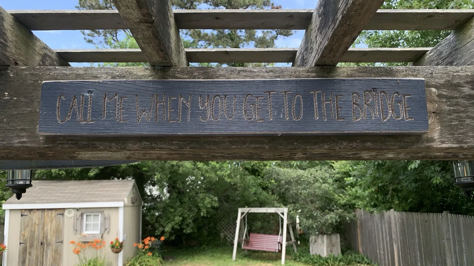 A carved wooden sign reading CALL ME WHEN YOU GET TO THE BRIDGE mounted on a pergola over a green backyard with a shed and swing.