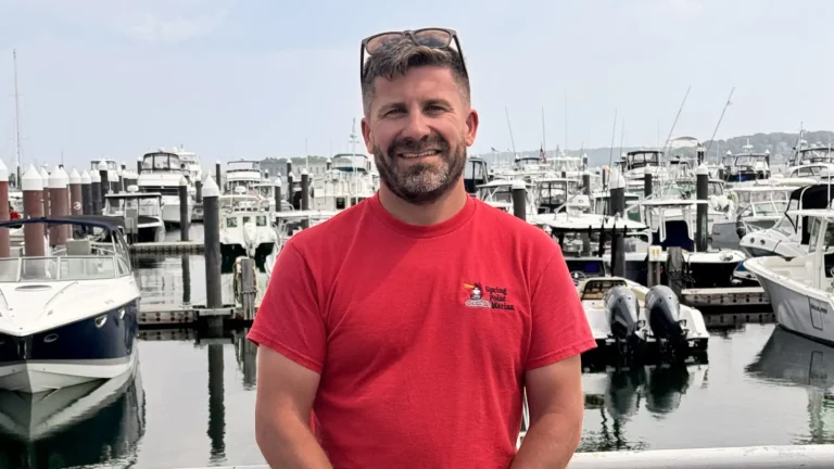 Peter Rauscher, founder of Marina Car, standing on a dock in a red Spring Point Marina shirt with rows of powerboats behind him in South Portland, Maine