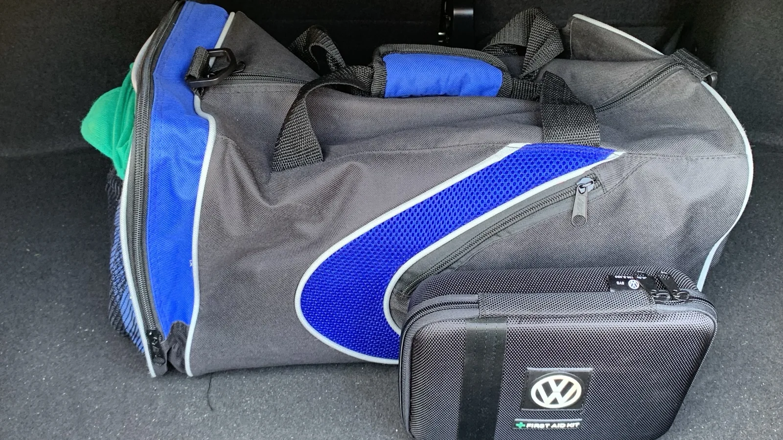 A gray and blue duffel bag and a black Volkswagen first aid kit sitting in the carpeted trunk of a car.