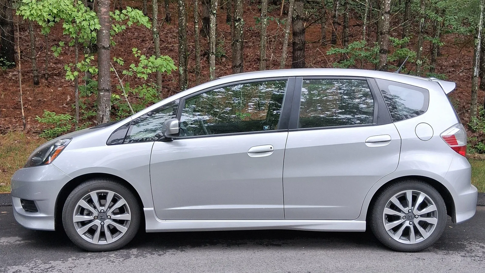 Side view of a silver 2013 Honda Fit hatchback parked beside a tree-lined rural road