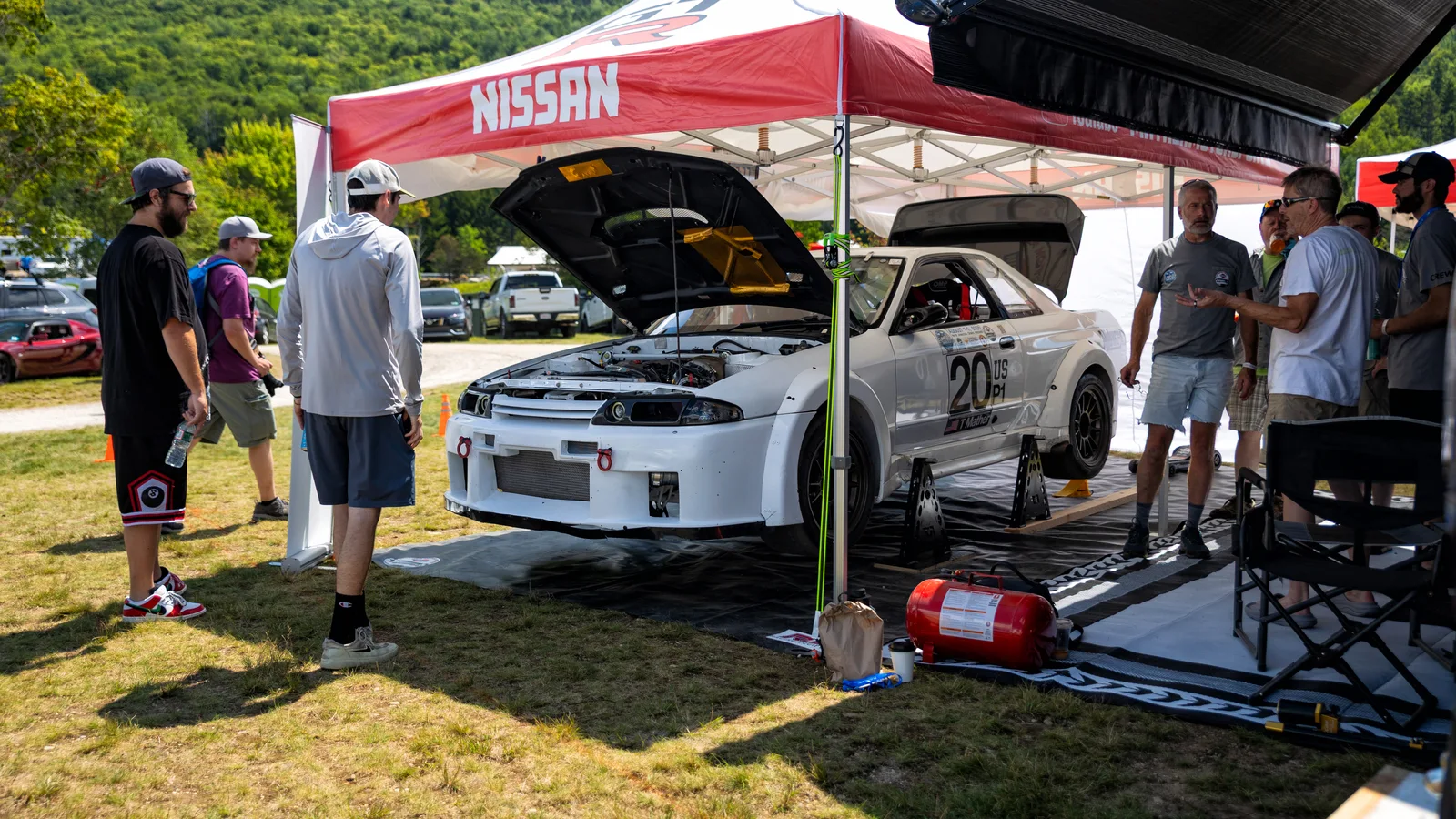 Spectators standing around a white Nissan Skyline GT-R race car with its hood up under a red Nissan team canopy in a grass paddock with green mountains behind.