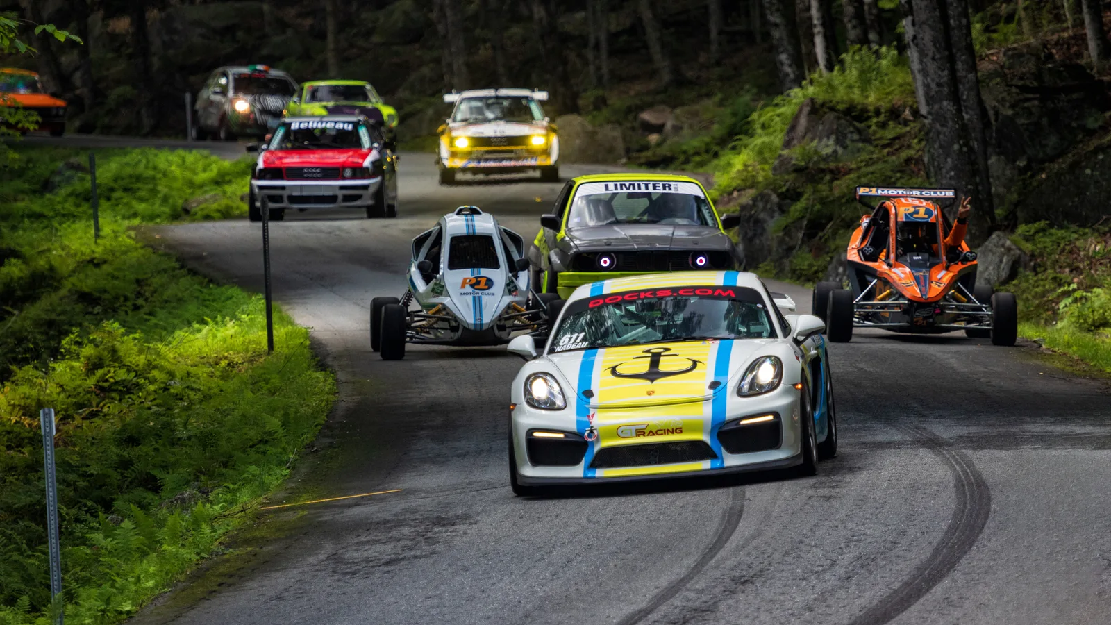 Several race cars and open-wheel crosskart buggies driving in single file down a curving section of the Mount Washington Auto Road surrounded by green forest.