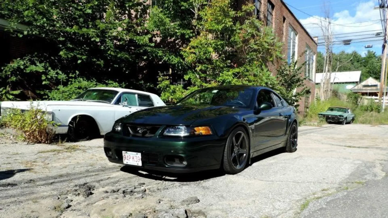 Dark green Ford Mustang parked on cracked pavement outside the abandoned brick South Weare Garage building in Weare, New Hampshire, with a white vintage sedan behind it
