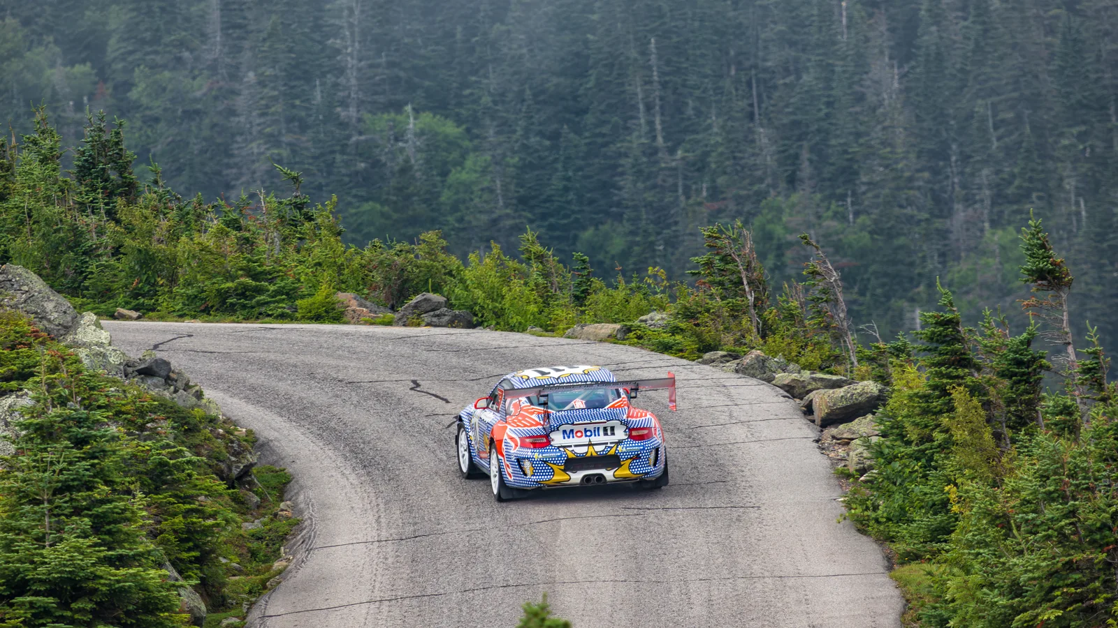 Porsche 911 race car in a red, white, and blue pop-art livery with Mobil 1 branding accelerating up a bend on the tree-lined Mount Washington Auto Road.