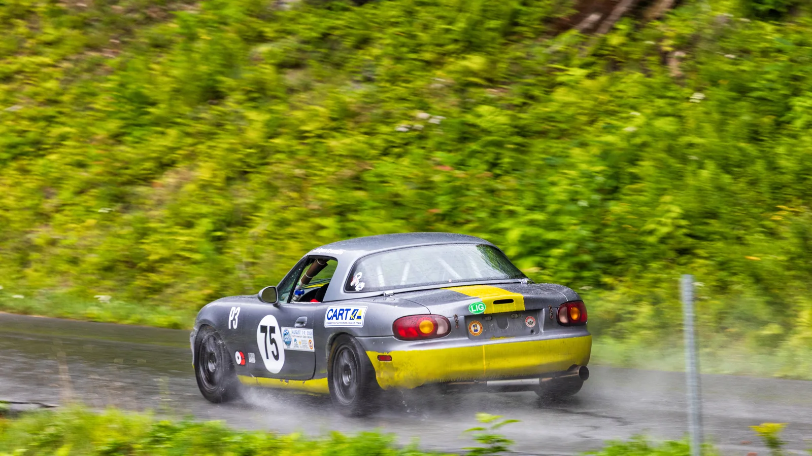 Grey and yellow Mazda MX-5 Miata race car number 75 spraying water off wet pavement on the Mount Washington Auto Road.