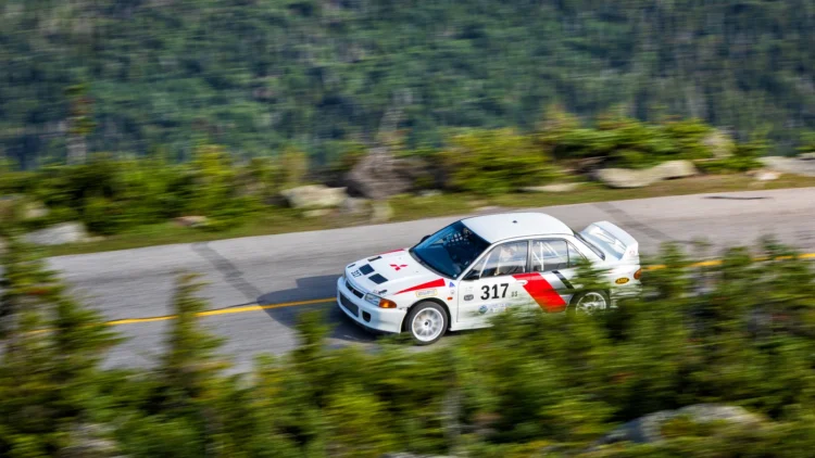White 1994 Mitsubishi Lancer Evolution II race car number 317 climbing a guardrail-free section of the Mount Washington Auto Road in New Hampshire, with evergreens and mountainside behind it.