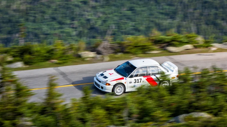 White 1994 Mitsubishi Lancer Evolution II race car number 317 climbing a guardrail-free section of the Mount Washington Auto Road in New Hampshire, with evergreens and mountainside behind it.