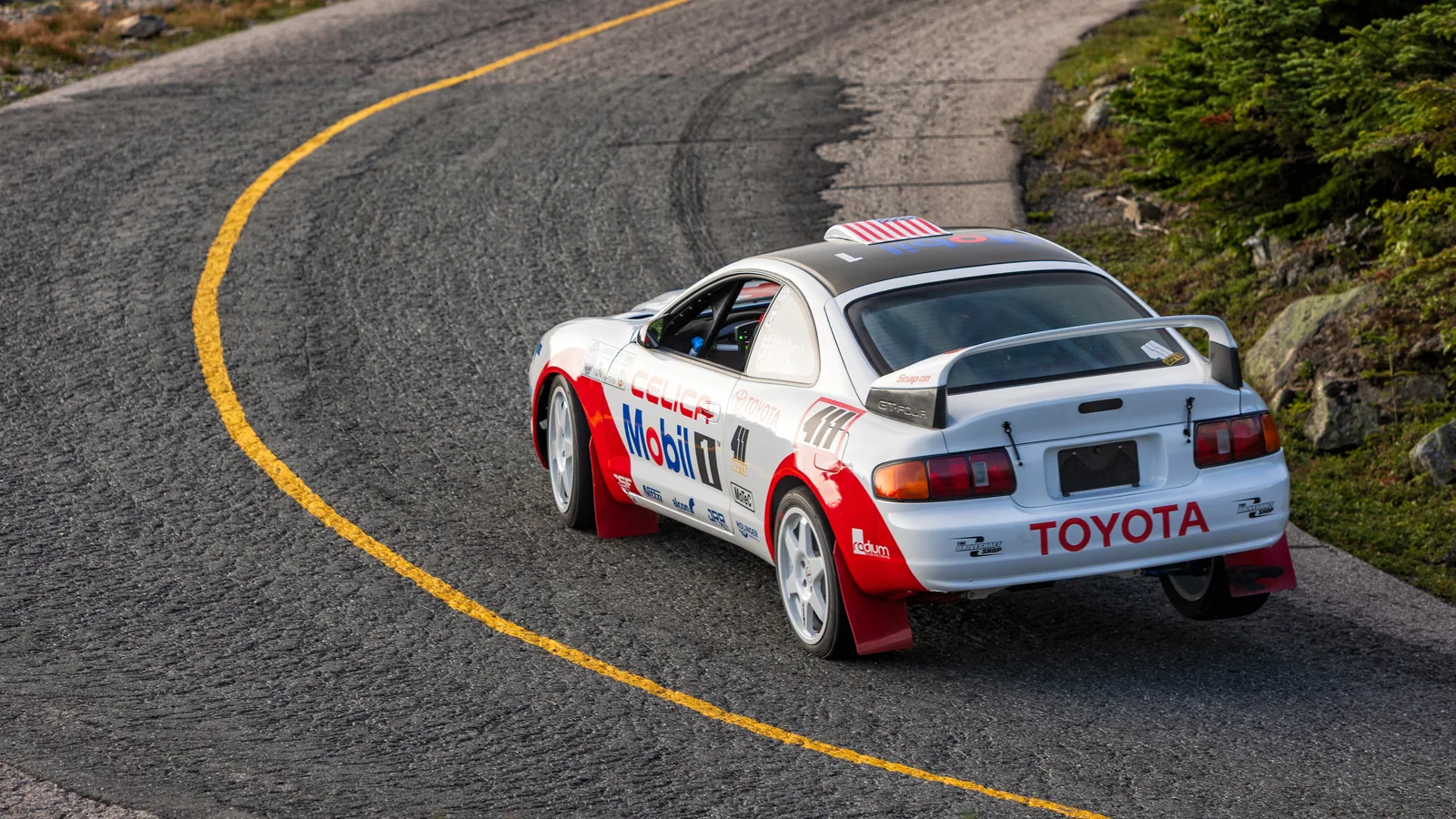 White and red Toyota Celica hillclimb car with Mobil 1 livery cornering on the Mount Washington Auto Road, seen from above and behind.