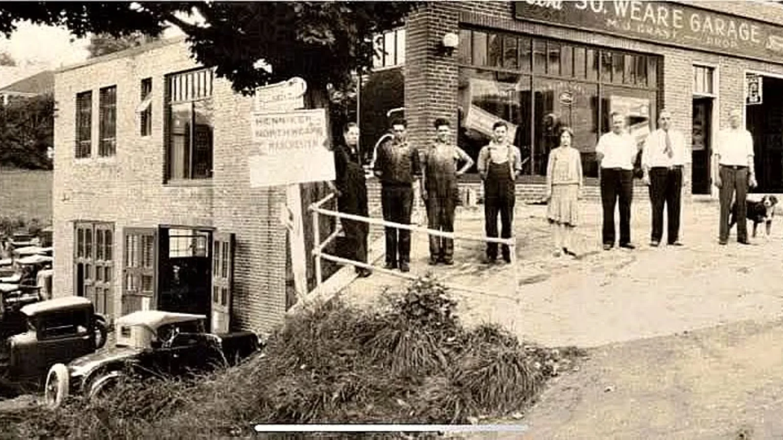 Sepia photo of South Weare Garage employees standing in a line outside the brick Ford dealership in Weare, New Hampshire, with vintage cars parked nearby