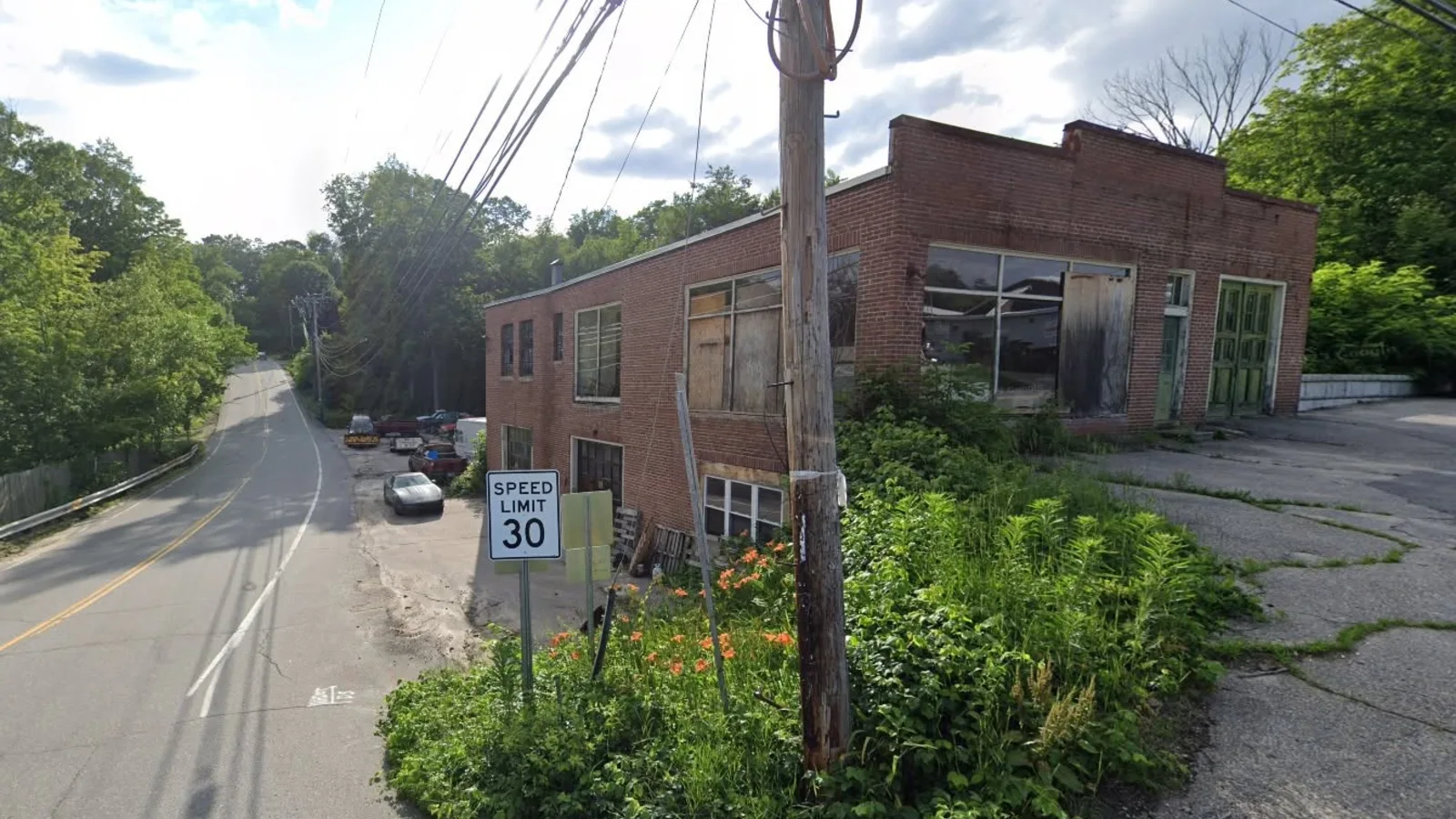 Google Street View of the weathered brick former South Weare Garage building at the corner of Route 114 and Short Street in Weare, New Hampshire, surrounded by overgrown brush