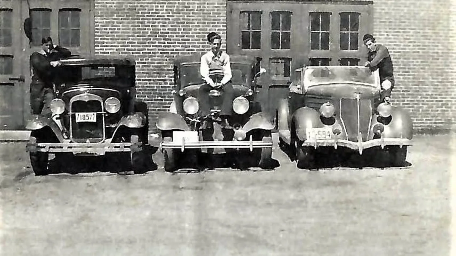 Black and white photo of three young men posing with three 1930s Ford automobiles in front of a brick garage building in Weare, New Hampshire