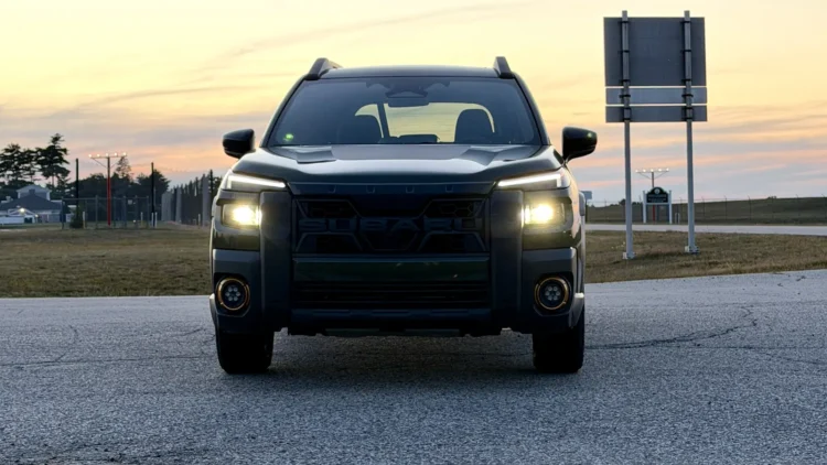 Head-on view of a dark green 2026 Subaru Outback Wilderness with LED headlights and round fog lights glowing, parked on cracked pavement under a pastel dusk sky
