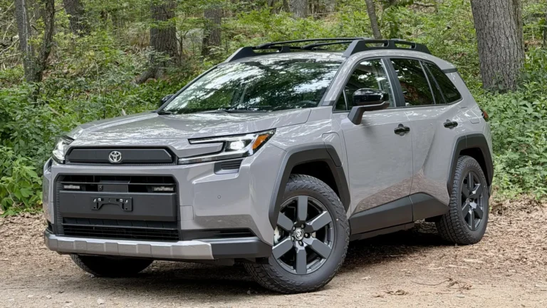 Gray 2026 Toyota RAV4 Plug-in Hybrid Woodland parked on gravel in front of tall trees, seen from the front three-quarter angle