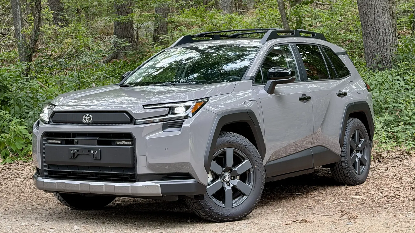 Gray 2026 Toyota RAV4 Plug-in Hybrid Woodland parked on gravel in front of tall trees, seen from the front three-quarter angle