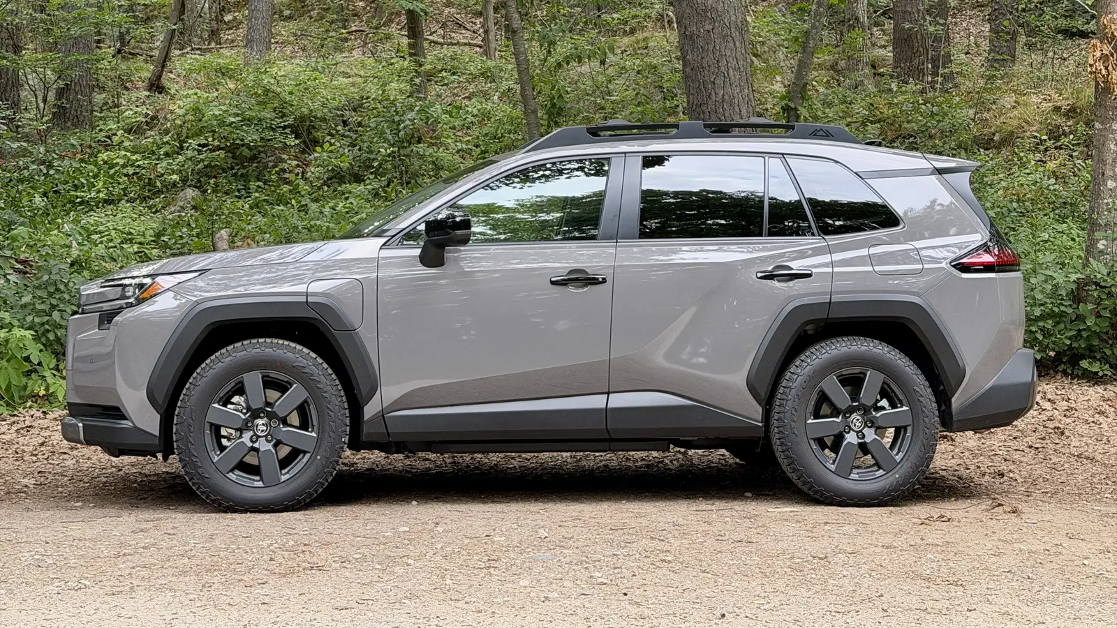 Side profile of a gray 2026 Toyota RAV4 Plug-in Hybrid Woodland on a gravel pull-off beside a forest