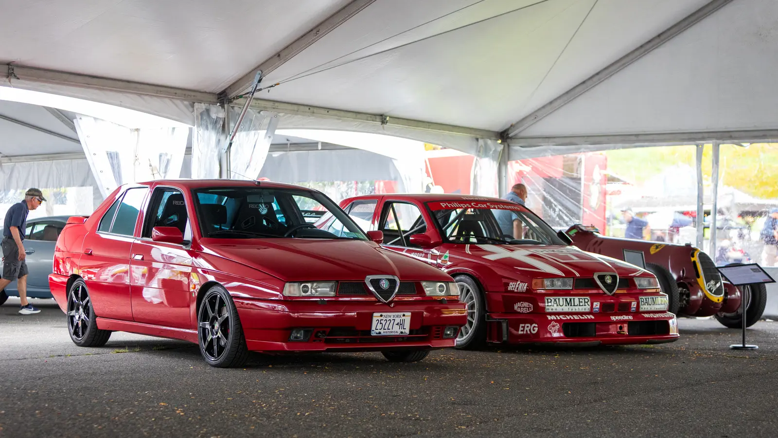 A red Alfa Romeo 155 sedan with black wheels parked under a white tent beside a red Alfa Romeo 155 race car covered in sponsor decals, with a prewar Alfa Romeo grand prix car behind them.