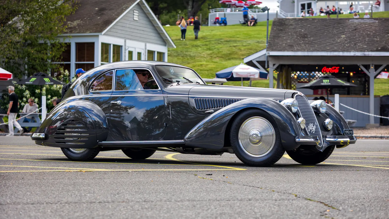 A dark grey prewar Alfa Romeo 8C 2900B Berlinetta coupe with flowing fenders and a tall shield grille driving slowly past spectators at a racetrack paddock.