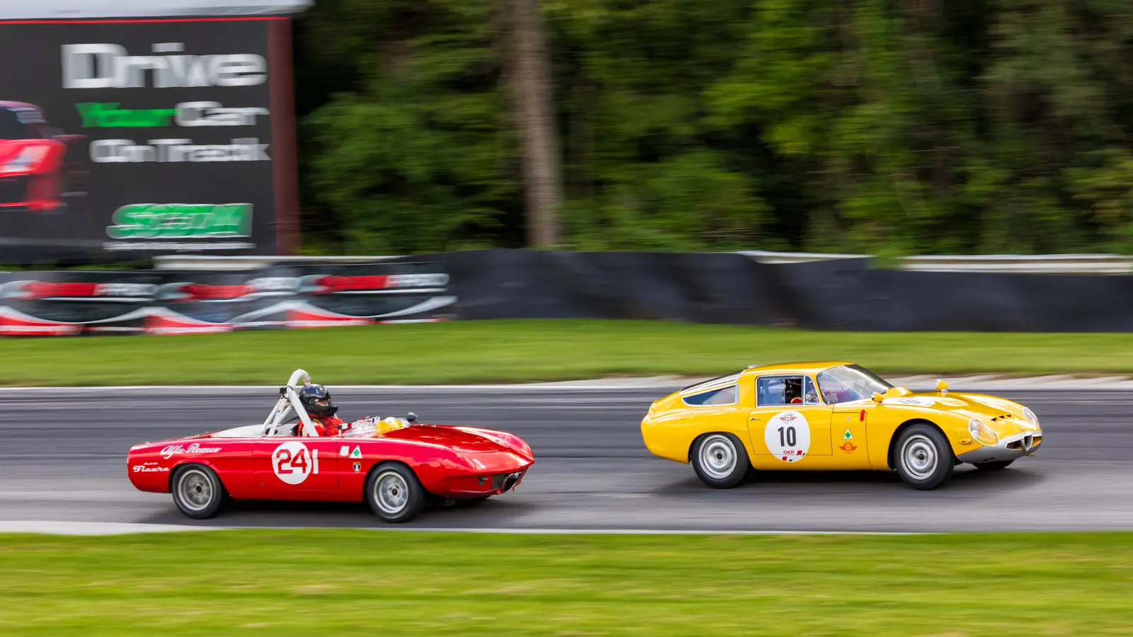 A yellow Alfa Romeo Giulia TZ coupe running alongside a red open-top Alfa Romeo race car on a racetrack, both panned against blurred trackside signage.