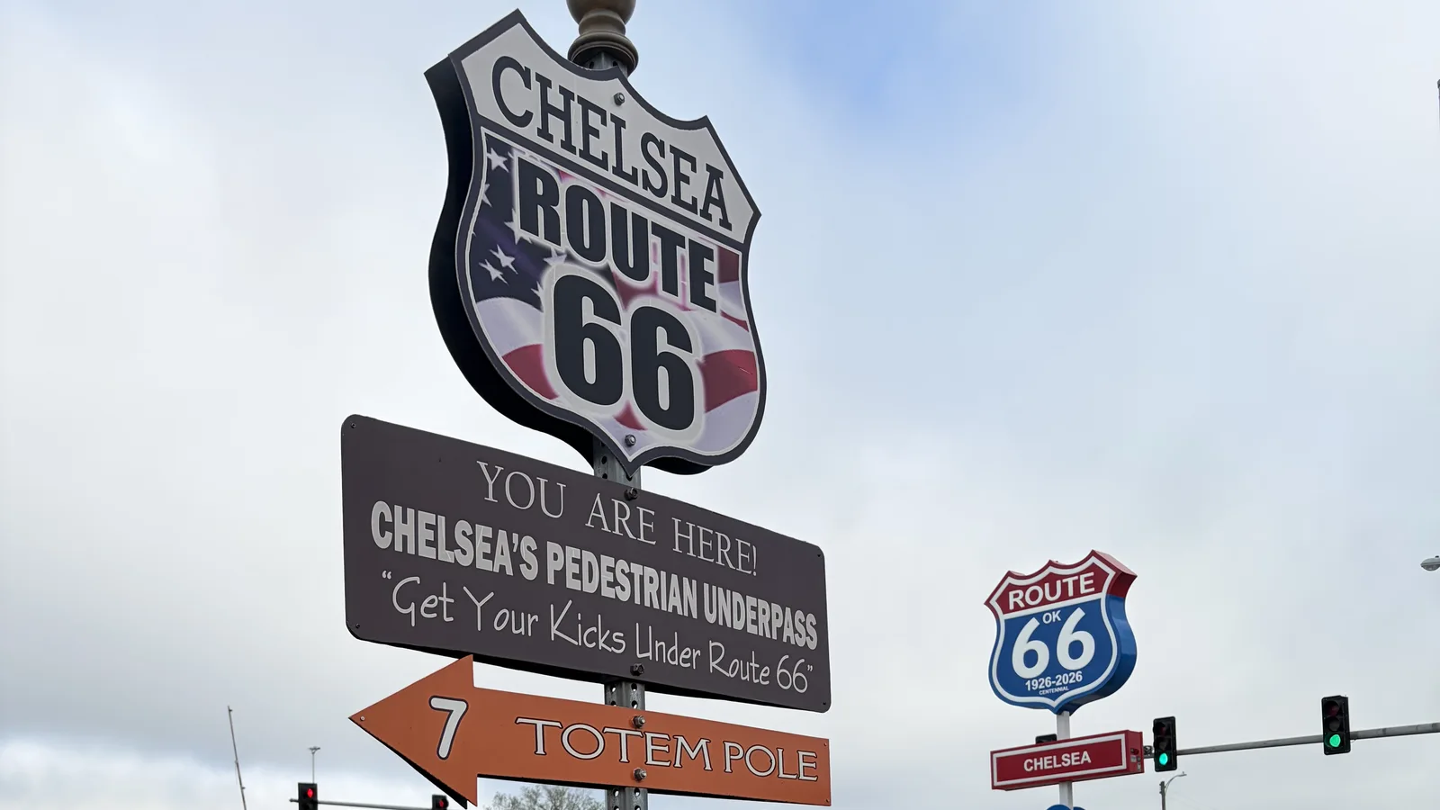 A Chelsea Route 66 shield atop a signpost reading You Are Here, Chelsea's Pedestrian Underpass, with an arrow pointing 7 miles to the Totem Pole, beside a blue Oklahoma Route 66 1926-2026 centennial shield.