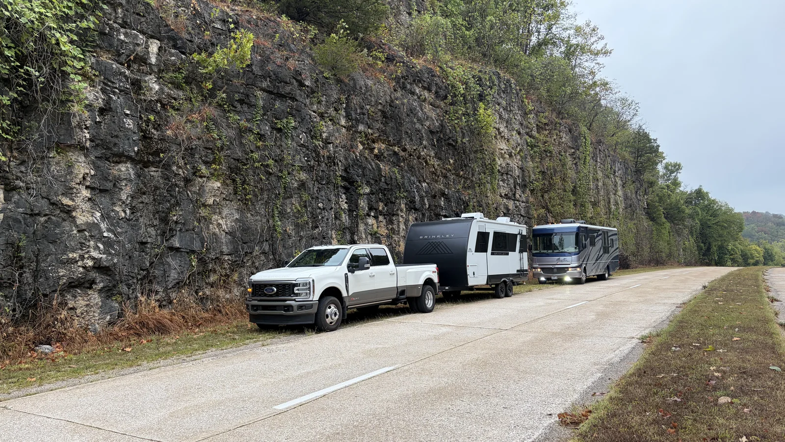 White Ford F-350 Super Duty dually pickup towing a Brinkley travel trailer, parked ahead of a motorhome on a concrete four-lane section of Route 66 beside a limestone rock cut near Newburg, Missouri.