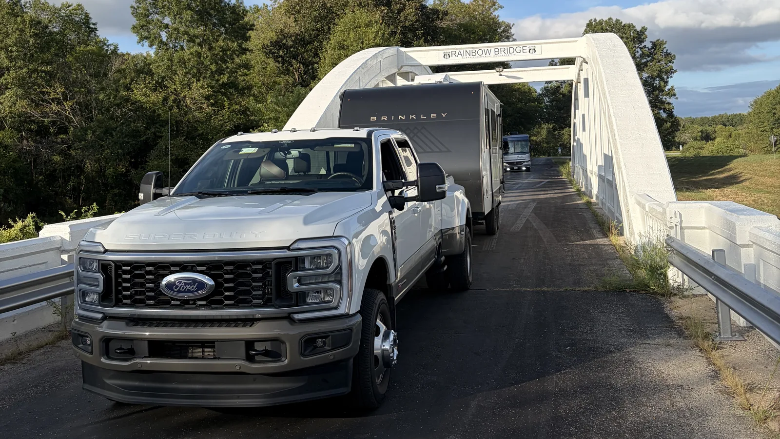 White Ford Super Duty dually pickup towing a Brinkley travel trailer across the white concrete arch of the Rainbow Bridge on Route 66 in Kansas, followed by a motorhome.