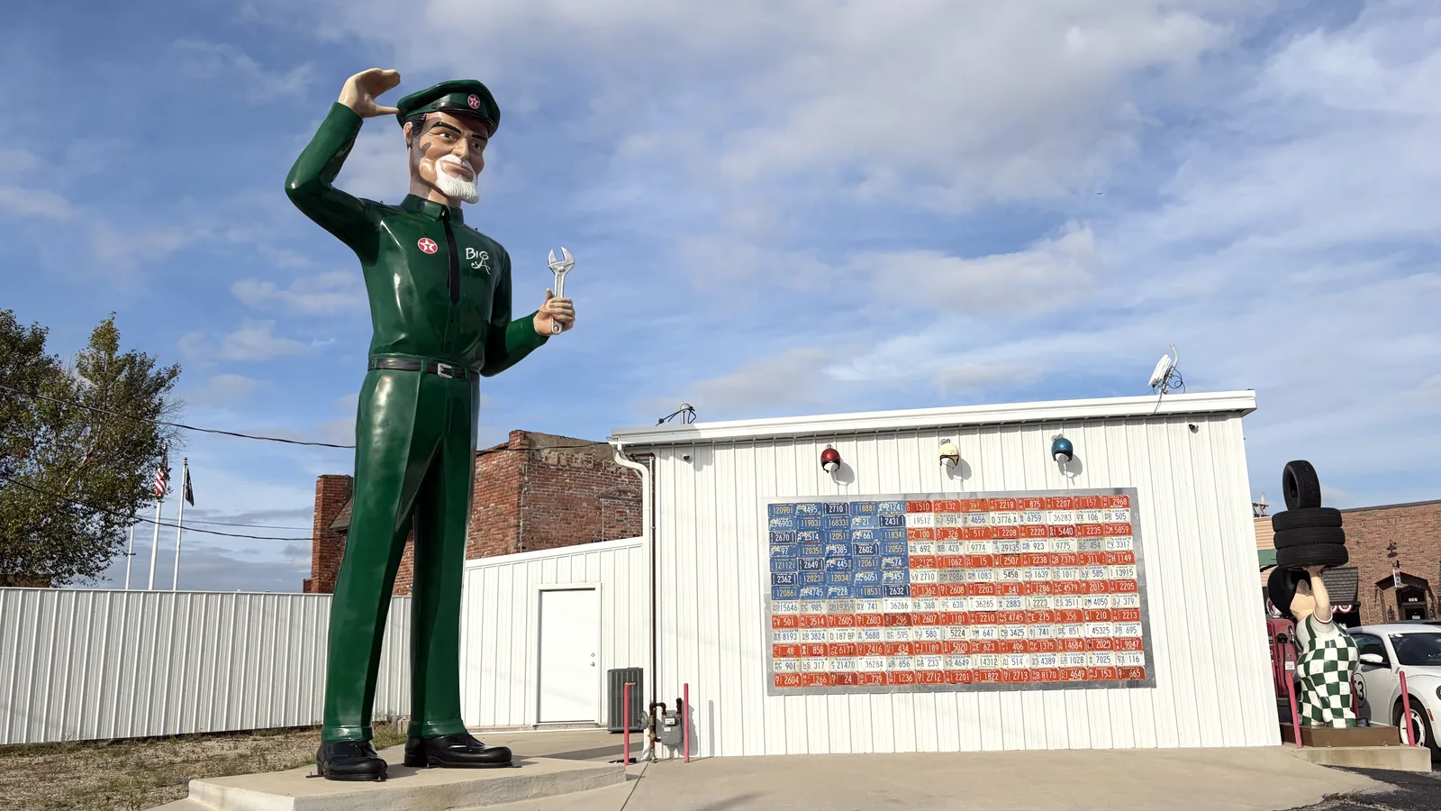 A giant service station attendant statue in a green uniform saluting and holding a wrench, beside an American flag made of license plates on a white building along Route 66 in Galena, Kansas.