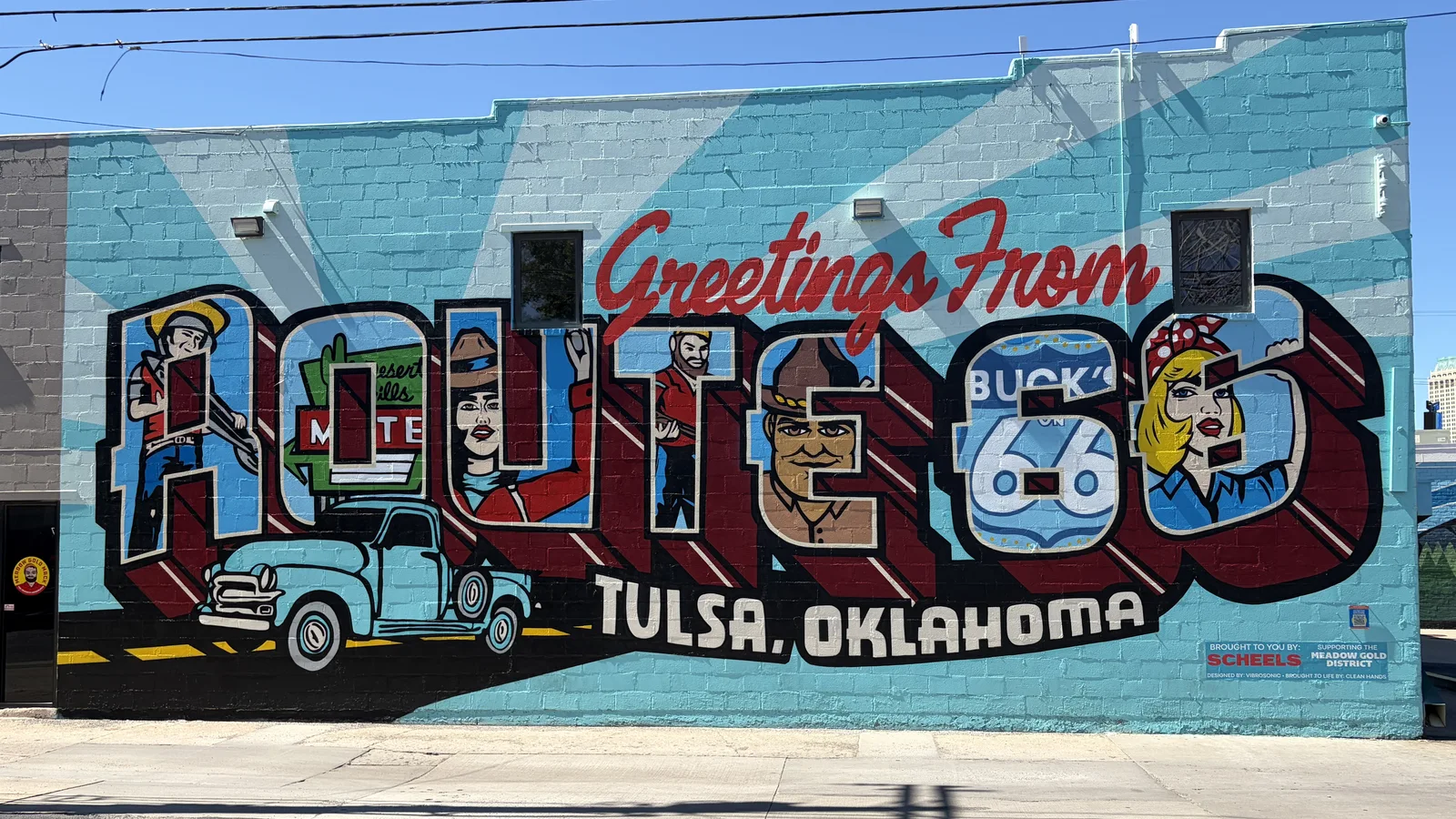 A large postcard-style Greetings From Route 66, Tulsa, Oklahoma mural painted on a brick wall, featuring roadside giant statues and a vintage pickup truck.