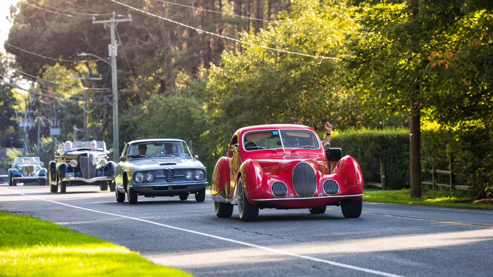 A red streamlined prewar coupe leads a silver Alfa Romeo coupe and two 1930s convertibles down a two-lane road in evening light, the lead driver waving an arm out the window.
