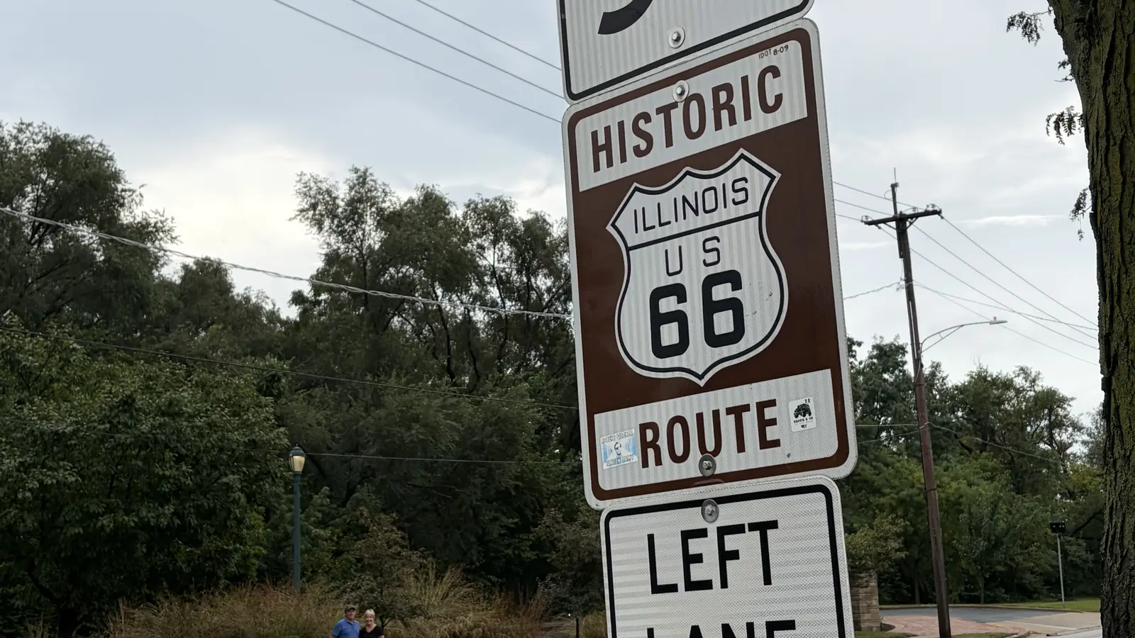 Historic Illinois US Route 66 brown marker mounted on a signpost below an Illinois Route 53 South sign along a tree-lined road
