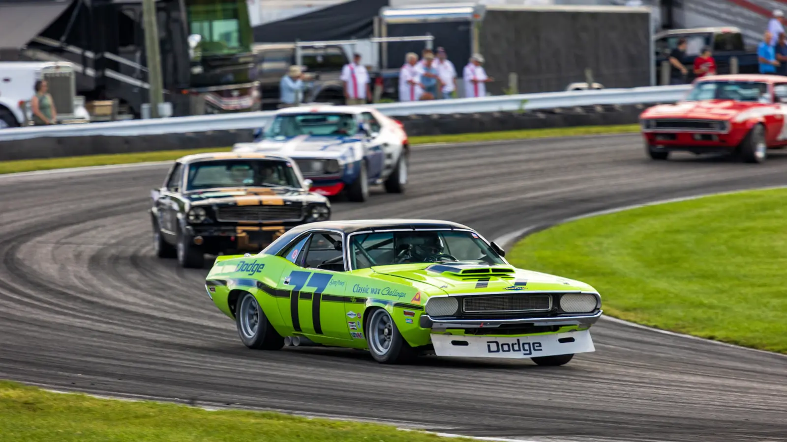 A bright green Dodge Challenger race car cornering ahead of a dark Ford Mustang and a red Chevrolet Camaro on a racetrack, with motorhomes and spectators lining the fence behind.