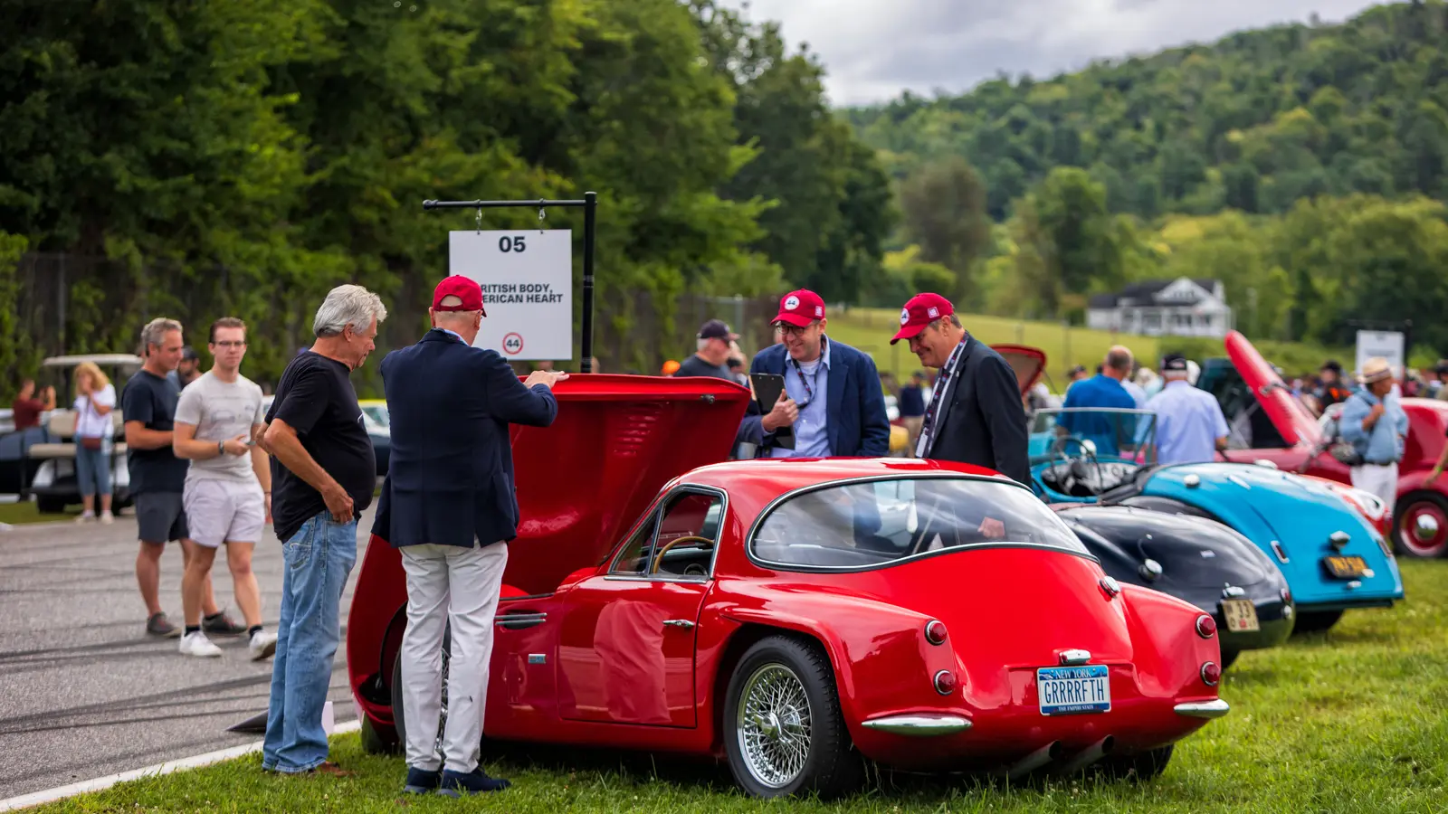 Two judges in red caps and blazers inspect a red TVR Griffith with its hood open while the owner and another man talk beside it on a grass show field.
