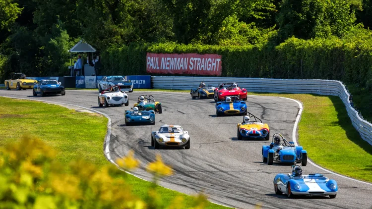 A pack of colorful vintage open-cockpit sports racing cars and small GT coupes sweeping down a tree-lined racetrack straight past a red banner reading Paul Newman Straight.