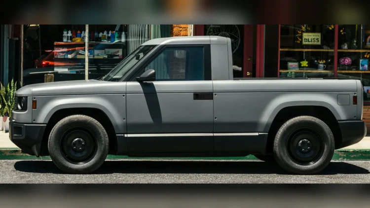 Matte gray Slate electric pickup truck with black steel wheels, parked at a curb in front of a storefront, seen in left side profile.