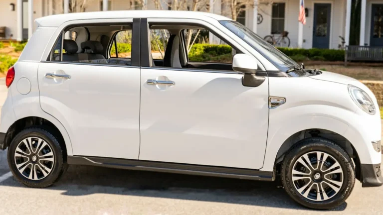 White Wink Mark3 electric microcar parked at a curb, seen in right side profile, in front of a white house with a bicycle on the porch.